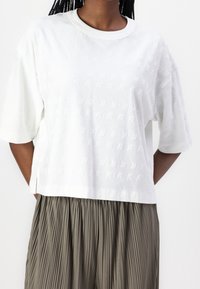 Femme portant un haut en tricot texturé blanc à manches courtes et une jupe plissée grise sur fond uni.