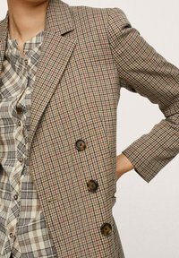 Veste blazer marron à carreaux, coupe ajustée, avec un col à revers cranté, trois boutons et un tissu texturé. Portée sur une chemise à carreaux.