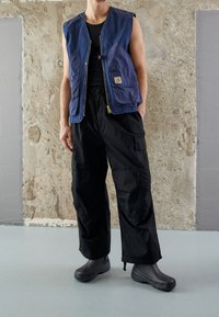 Granatowa bawełniana kamizelka bez rękawów z przednimi kieszeniami, czarne spodnie cargo i czarne klapki. Teksturowane tło dodaje kontrastu do stroju.