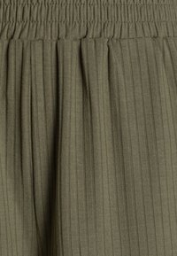 Tissu texturé de couleur vert olive, côtelé. Comprend une taille élastique et des plis doux pour un ajustement décontracté.