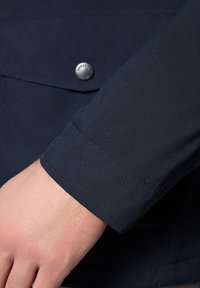 Morska niebieska rękaw kurtki wykonanej z gładkiego materiału, z metalowym zatrzaskiem w kolorze srebrnym i widocznymi detalami szycia. Całościowy design jest elegancki i dopasowany.