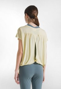 Kvinde med brunt hestehale iført en løs, lys gul kortærmet top over en blågrå tætsiddende skjorte og matchende leggings, set bagfra.