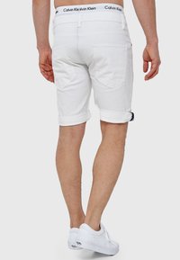 Shorts en denim blanc avec des poignets retroussés, comportant des poches arrière et une ceinture de marque Calvin Klein. Le modèle porte des baskets blanches.