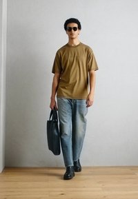Barna pamut póló rövid ujjakkal, világoskék farmernadrág, fekete bőrcsizma és fekete szövet táska. A modellen fekete napszemüveg van.