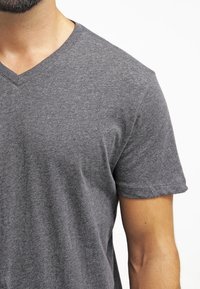 Uomo che indossa una semplice t-shirt grigia a maniche corte con scollatura a V, che mostra il petto e il braccio destro su uno sfondo bianco.