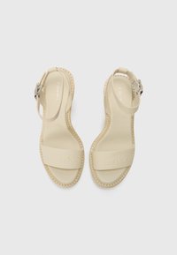 Beige sandaler med to brede stropper, som har preget merkevare. Justerbar ankelrem med sølvfarget maskinvare, gummisåle og glatt tekstur.