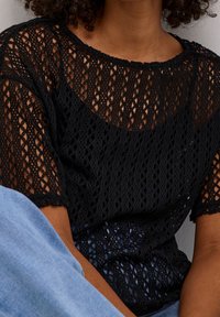 Top in pizzo nero con un design a maglia aperta, scollatura ampia e maniche corte, abbinato a denim blu chiaro. Tessuto leggero e strutturato.