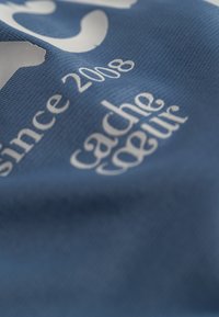 Blauwe stof met witte tekst, met een licht gestructureerd oppervlak waarop "sinds 2008" en "cache cœur" in elegante lettertypes is weergegeven.