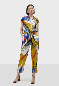 Pestrý overal v saténovom vzhľade; má golierový výstrih, krátke rukávy a abstraktný vzor modrej, červenej, zelenej a žltej farby.