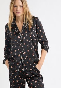 Czarny zestaw piżamowy w kwiaty wykonany z gładkiej tkaniny; zdobiony małymi różowymi kwiatami, białymi akcentami i guzikami. Długie rękawy z podwiniętymi mankietami.