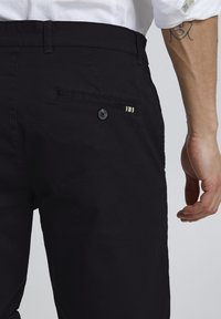 Schwarze Baumwollhosen mit einer geknöpften Gesäßtasche, die drei dekorative Streifen aufweist und eine glatte Textur hat.