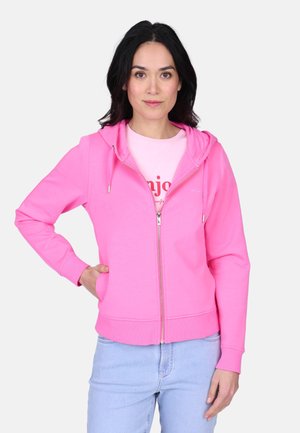 Femme aux cheveux foncés portant un sweat à capuche rose vif zippé sur un t-shirt rose clair et un jean bleu clair, debout avec une main dans la poche.