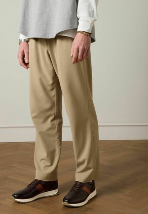 Man draagt beige broek, donderbruine sneakers met witte zolen en een grijs vest over een wit shirt, staande op een houten vloer.