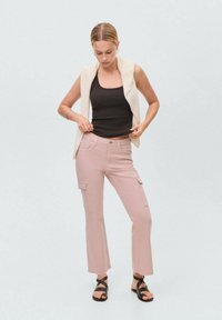 Roze cargobroek met platte zakken, gecombineerd met een zwarte tanktop en een crèmekleurige mouwloze jas. Strappy zwarte sandalen maken de outfit compleet.