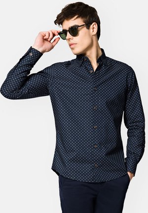 Granatowa koszula zapinana na guziki z małymi geometrycznymi wzorami, długie rękawy i brązowe guziki. Połączona z ciemnymi spodniami i czarnymi okularami przeciwsłonecznymi.