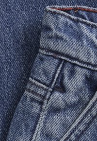 Zbliżenie na niebieską tkaninę denimową, ukazujące szczegółowe przeszycia oraz złożony obszar pasa z widoczną pętelką na pasek.