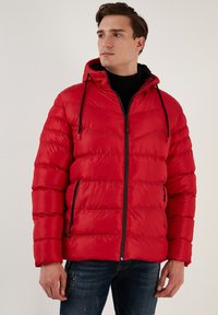 Rød pufferjakke med hætte, der har en quiltet tekstur, en frontlynlås og sorte detaljer. Båret over en sort turtleneck for kontrast.