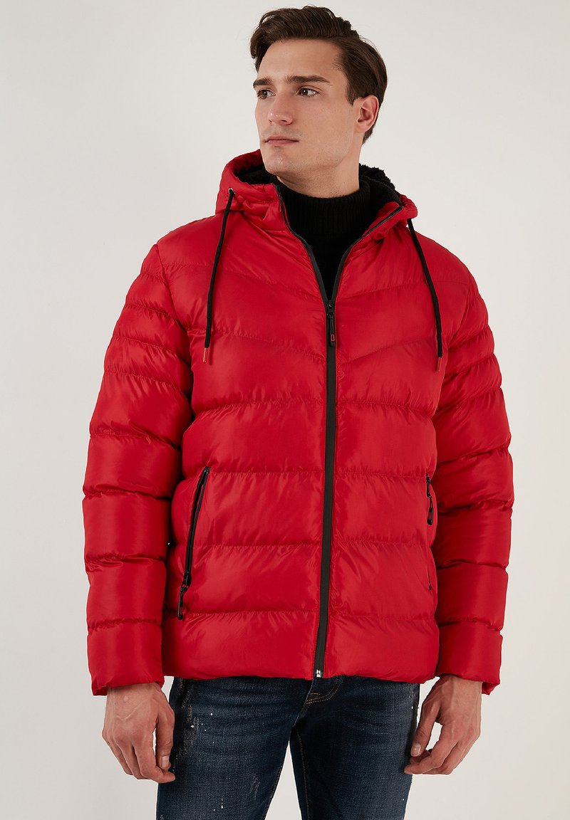 Rød pufferjakke med hætte, der har en quiltet tekstur, en frontlynlås og sorte detaljer. Båret over en sort turtleneck for kontrast.