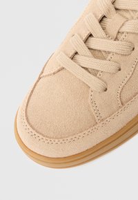 Sneaker beige in suede con lacci spessi beige, cuciture dettagliate e suola in gomma marrone chiaro su uno sfondo bianco.