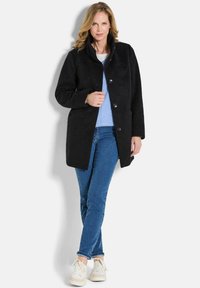 Cappotto nero con bottoni e colletto alto, realizzato in tessuto testurizzato. Indossato sopra una maglietta azzurro chiaro e abbinato a jeans e sneakers bianche.