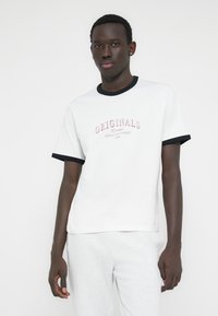 Biele bavlnené tričko s čiernym rebrovaným výstrihom a rukávmi. Obsahuje červený nápis „ORIGINALS“ s grafikou pod ním. Nosené so sivými teplákmi.