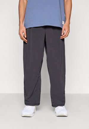Homme portant un pantalon noir ample, une chemise bleue et des baskets blanches, debout devant un fond clair uni.