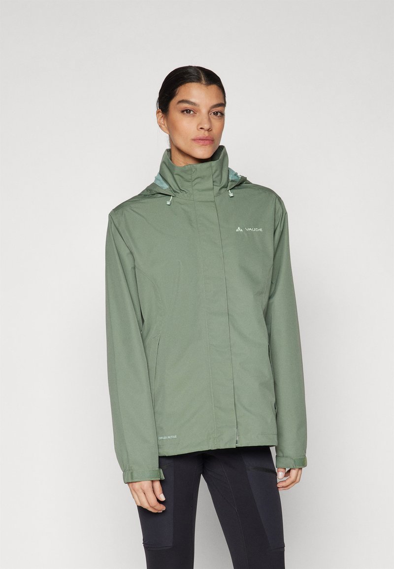 Femme portant une veste imperméable vert clair à col montant et capuche, debout les mains détendues le long du corps, fond blanc neutre.
