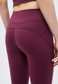 Γυναίκα που φοράει ψηλόμεσα, σκούρα μωβ κολάν με ορατή ραφή στο πίσω και πλαϊνό μέρος, στέκεται με το χέρι χαλαρωμένο στο πλάι.