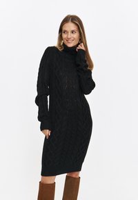 Dzianinowa czarna sukienka z wysokim kołnierzem, ozdobiona wzorem w warkocze, z prążkowanymi mankietami i dołem. Noszona z brązowymi kozakami do kolan. Miękka w dotyku.