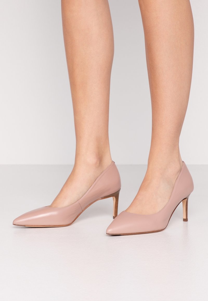Femme portant des escarpins en cuir rose nude à bout pointu avec talons aiguilles de hauteur moyenne sur un sol blanc.