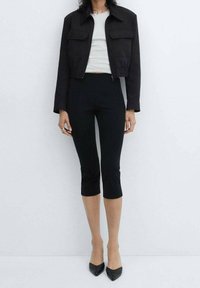 Femme portant une veste noire courte, un pantalon noir court, un haut blanc, des talons pointus noirs, debout contre un mur clair uni.