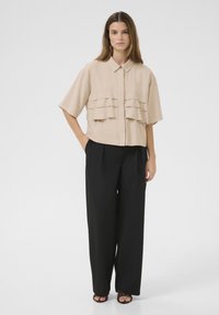Short-sleeve beige shirt with layered ruffles on the front, paired with black wide-leg trousers, worn by a model standing against a white background.