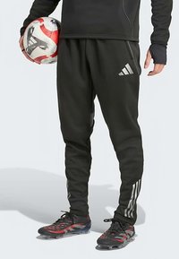 Osoba v černém sportovním oblečení Adidas drží fotbalový míč červené, bílé a černé barvy, má na sobě černé a červené kopačky se zavázanými tkaničkami.