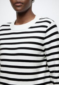 Gros plan d'une personne portant un pull blanc avec des rayures horizontales noires et une petite boucle d'oreille argentée visible sur une oreille.