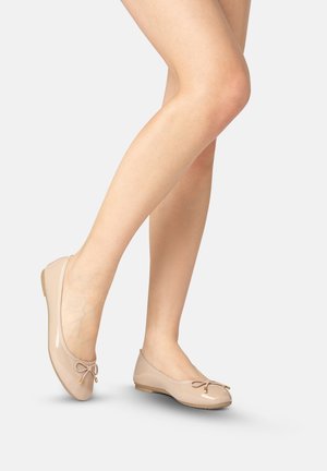 Piernas con bailarinas de charol color nude con pequeños lazos sobre un fondo gris claro, un pie ligeramente levantado.