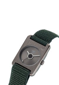 Montre carrée avec un boîtier gris métallisé, un bracelet tissé vert et un cadran minimaliste avec des aiguilles argentées et un logo Adidas.