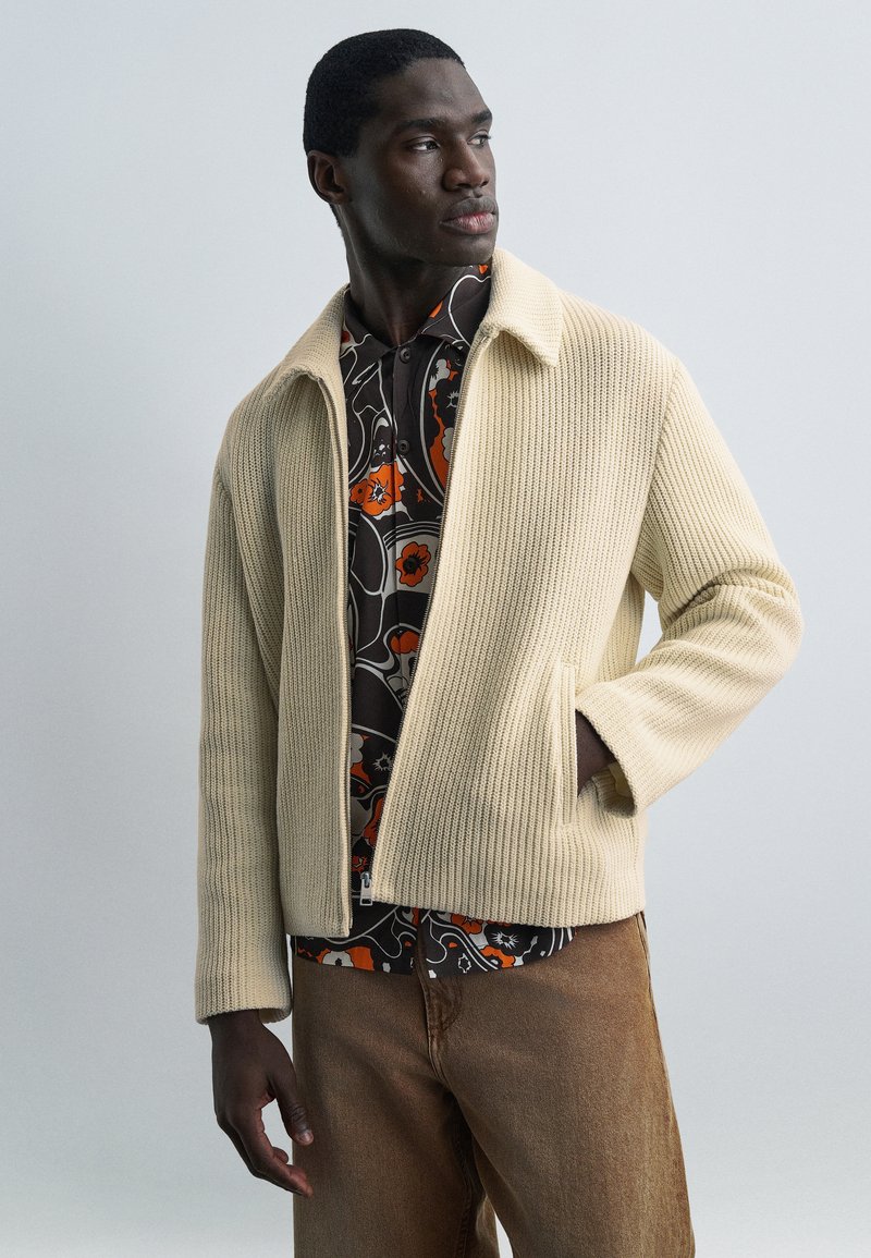 Άνδρας που φοράει μπεζ πλεκτό σακά κι πάνω από μαύρο και πορτοκαλί πουκάμισο με σχέδια, καφέ παντελόνια, στέκεται με το ένα χέρι στην τσέπη του σακακιού, κοιτάζει δεξιά.