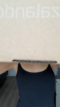 Caixa de cartão marrom segurada acima, com uma textura suave e uma marcação sutil. O fundo inclui um piso de madeira clara e móveis.