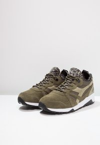 Oliwkowo-zielone buty sportowe wykonane z zamszu i siateczki, posiadające wzorzyste wykończenie wnętrza, czarną gumową podeszwę oraz kontrastującą białą podeszwę środkową.