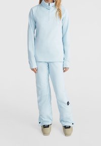 Kobieta w jasnoniebieskich spodniach narciarskich i dopasowanej kurtce zapinanej na zamek, stojąca z rękami swobodnie wzdłuż ciała na białym tle.