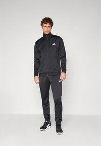 Fato de treino atlético preto feito de tecido liso, com uma jaqueta com fecho de correr e colarinho alto, e calças combinando, ambos com detalhes em branco do logótipo.