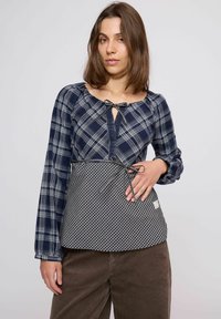 Tengerészkék kockás hosszú ujjú ing zsinóros kulcslyuk nyakkal és kockás mintájú derékrésszel, barna kordnadrág fölött viselve.