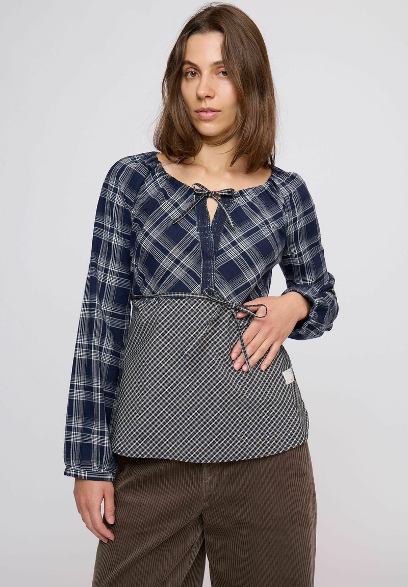 Tengerészkék kockás hosszú ujjú ing zsinóros kulcslyuk nyakkal és kockás mintájú derékrésszel, barna kordnadrág fölött viselve.