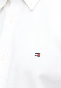 Λευκό πουκάμισο με γιακά και μικρό κεντημένο ορθογώνιο λογότυπο σε ναυτικό μπλε, λευκό και κόκκινο στο στήθος, κουμπωμένο.