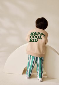 Kind, das sich auf einen weißen geschwungenen Block lehnt, trägt einen beigen Sweatshirt mit dem Aufdruck "HAPPY COOL KID", grün-weiß-blau gestreifte Hosen und weiße Sneaker.