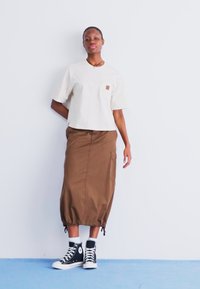 Beige cropped shirt met korte mouwen en een borstzak, gecombineerd met een bruine rok tot de enkels met zijzakken en een drawstringzoom, gedragen met zwarte sneakers.