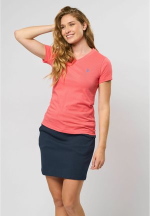 Korallenfarbene V-Ausschnitt-T-Shirt mit kurzen Ärmeln, kombiniert mit einem taillierten Marineblauen Rock. Aus Baumwollmaterial mit einem dezenten gestickten Logo auf der Brust.