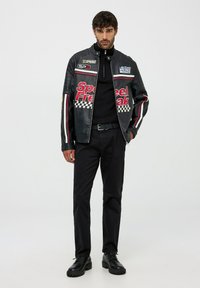 Giacca di pelle nera con testo rosso "Speed Freak", dettagli in pattern a scacchi e cerniera. Indossata sopra un maglione nero e abbinata a pantaloni neri e stivali.