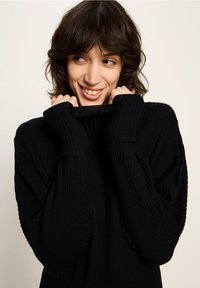 Schwarzer gerippter Strickpullover mit hohem Kragen und verlängerten Bündchen, lockere Passform und subtile Texturdetails.