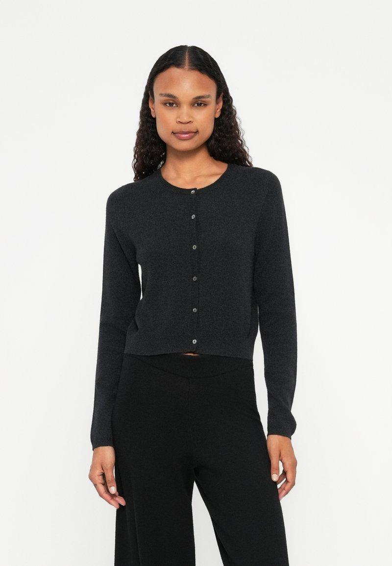 Μαύρο κοντό καρφίτσα φτιαγμένο από μαλακό πλέξη, με στρογγυλή λαιμόκοψη, μακριά μανίκια και κουμπί στην μπροστινή πλευρά.